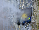 Ein Atemschutzgeräteträger kriecht durch einen Wanddurchbruch ins Freie. Dichter Rauch quillt aus der Öffnung hinter ihm.