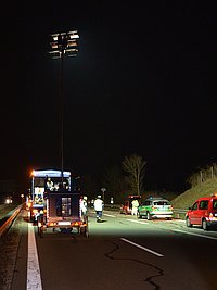 Eine nächtliche Unfallstelle auf einer Autobahn wird mit dem Lichtmastanhänger ausgeleuchtet. Fahrzeuge und Einsatzkräfte von Polizei und THW sind im Vordergrund.
